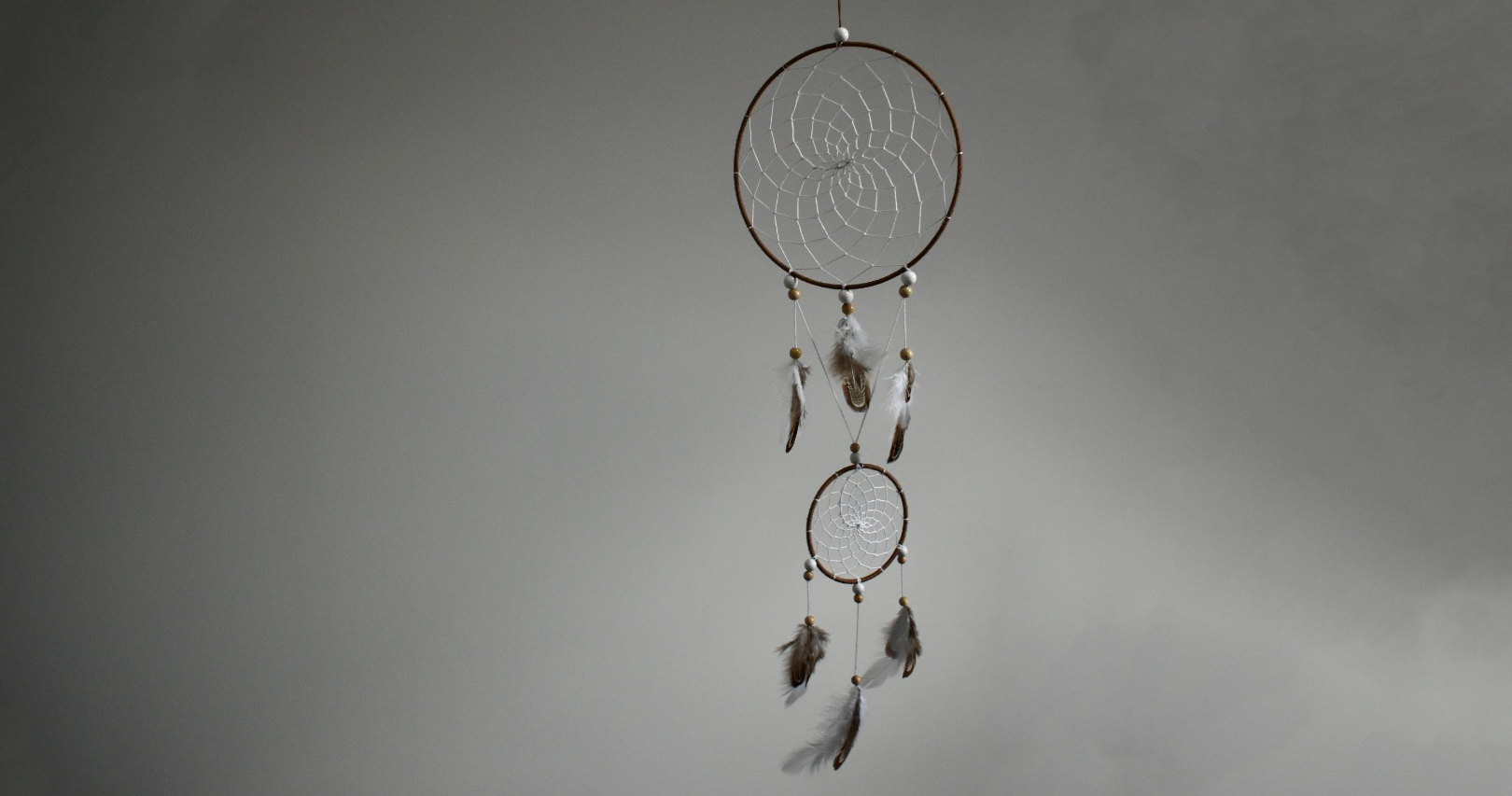 Attrape-rêve suspendu avec cercles bruns et blancs, perles bois clair et plumes naturelles sur fond blanc gris.